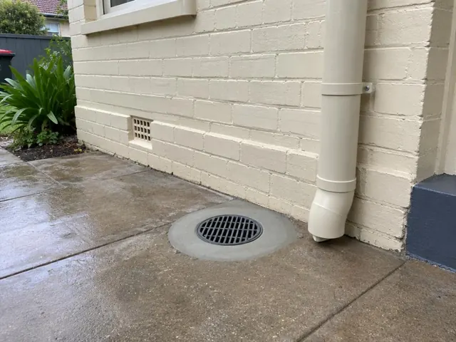 Outdoor drain grate beside a cream brick wall and downpipe on wet paved ground in Berala